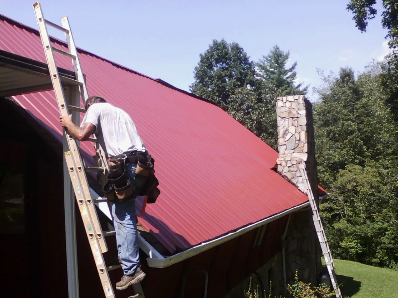 Roofer on a ladder beside a red metal roof and stone chimney — Soffit & Fascia Repair by Roofing Brothers in Verona